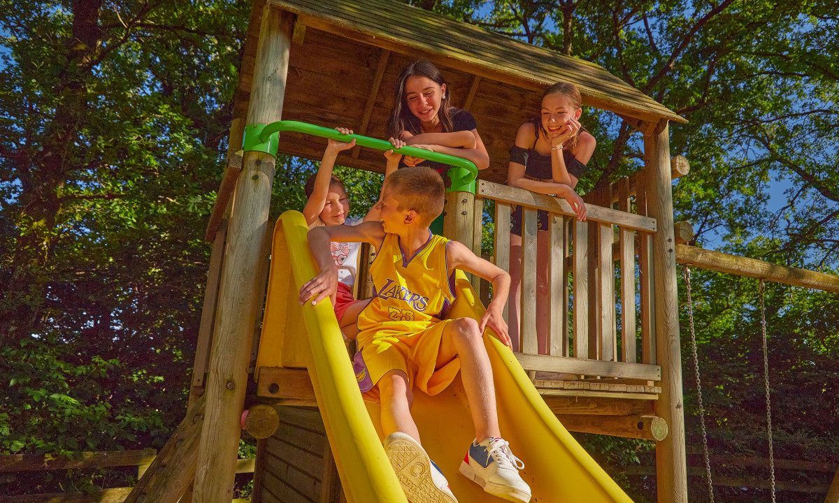 At Terrasses du Lac, a boy glides down a yellow slide while three friends observe from a wooden playhouse.
