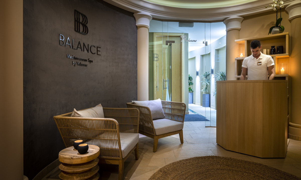 Reception desk with a person standing, alongside wicker chairs, in a welcoming spa lobby with soft lighting at Valamar Obertauern Hotel