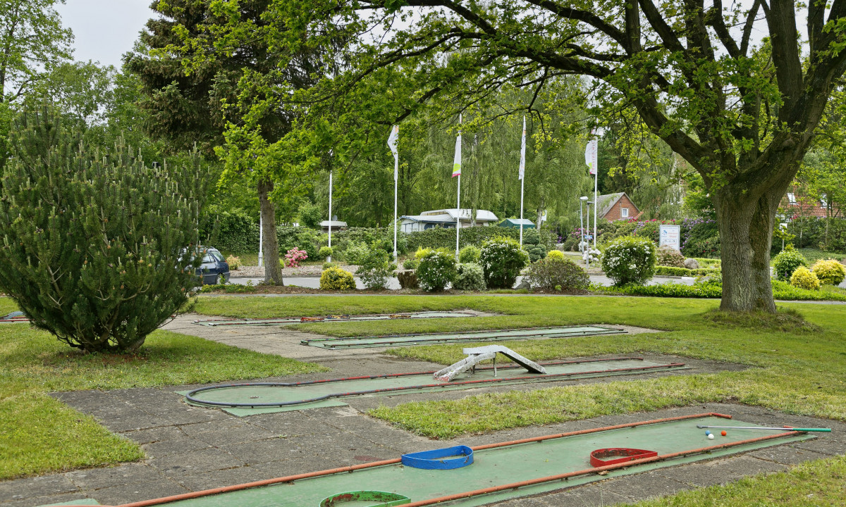 At KNAUS Campingpark Wingst, a miniature golf course sits idle amidst vibrant green trees and bushes, with flags and parked vehicles visible in the background.