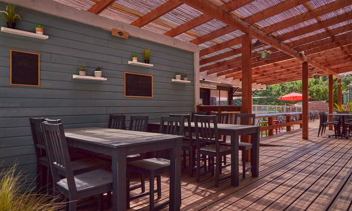 At Le Chêne Vert, wooden tables and chairs are positioned beneath a pergola; potted plants and chalkboards decorate the nearby walls; vibrant foliage encircles the area.
