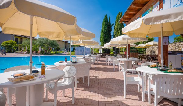 White patio tables with matching chairs under umbrellas, set near a pool, surrounded by greenery and buildings at Residence Eden