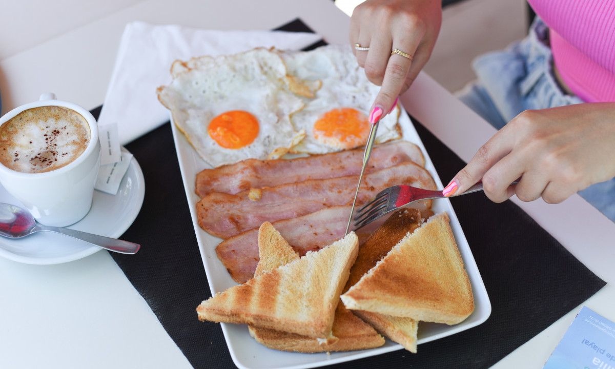 Person cutting toast and bacon, with fried eggs and coffee on a white table at Resort Els Pins