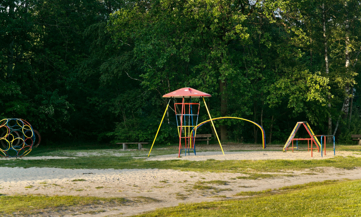 At KNAUS Campingpark Oyten, vibrant metal play structures stand unused in a sandy play zone, encircled by verdant trees.