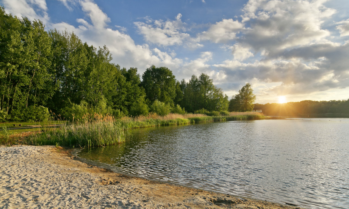 At KNAUS Campingpark Oyten, the sun descends over a tranquil lake, flanked by a sandy shore and dense foliage, with a cloudy sky in the background.