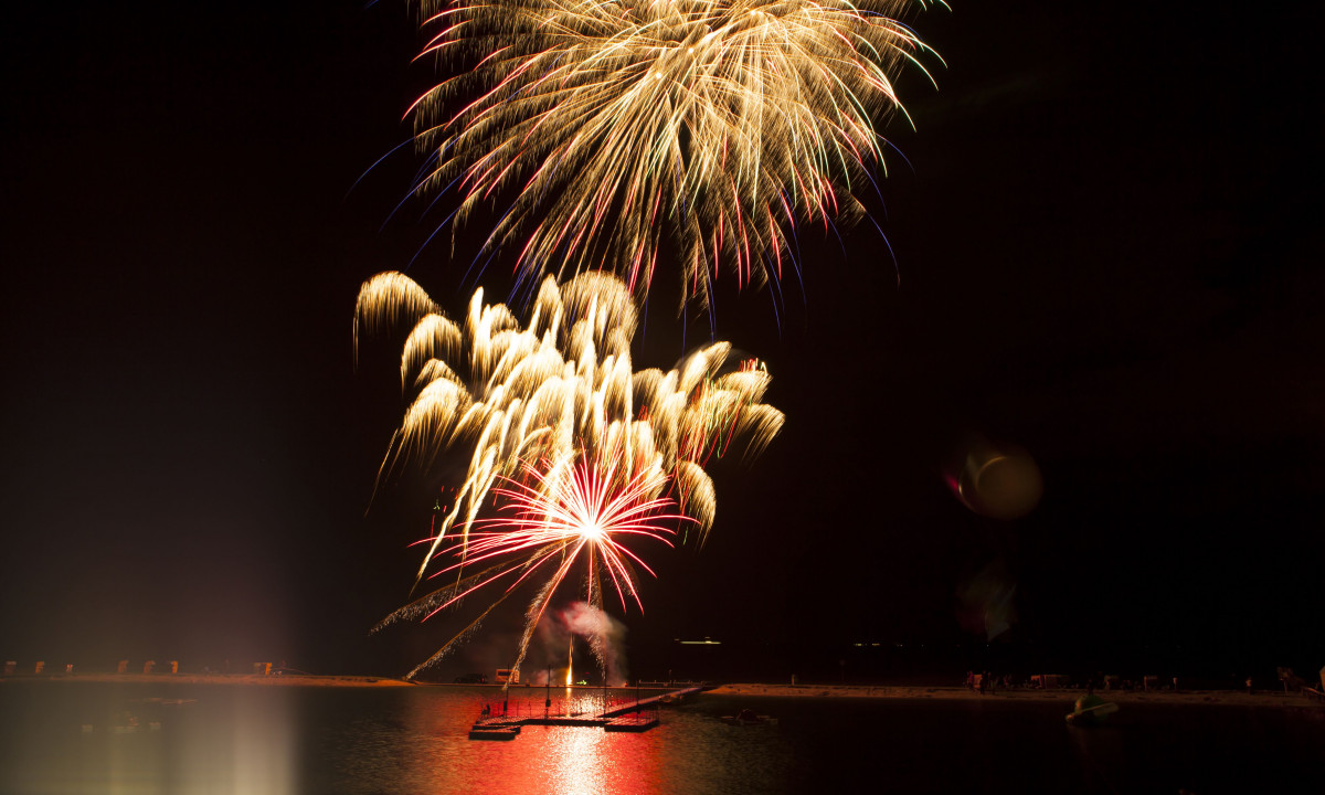 At KNAUS Campingpark Eckwarderhörne, pyrotechnics explode above a lakeside, casting shadows of swimmers in the water against the dark night sky.