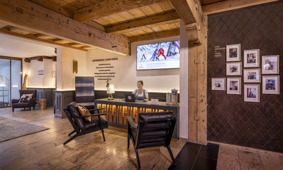 Receptionist sitting at a wooden desk, typing on a laptop, in a warmly lit, cozy hotel reception area at Kesselspitze Valamar Collection Hotel