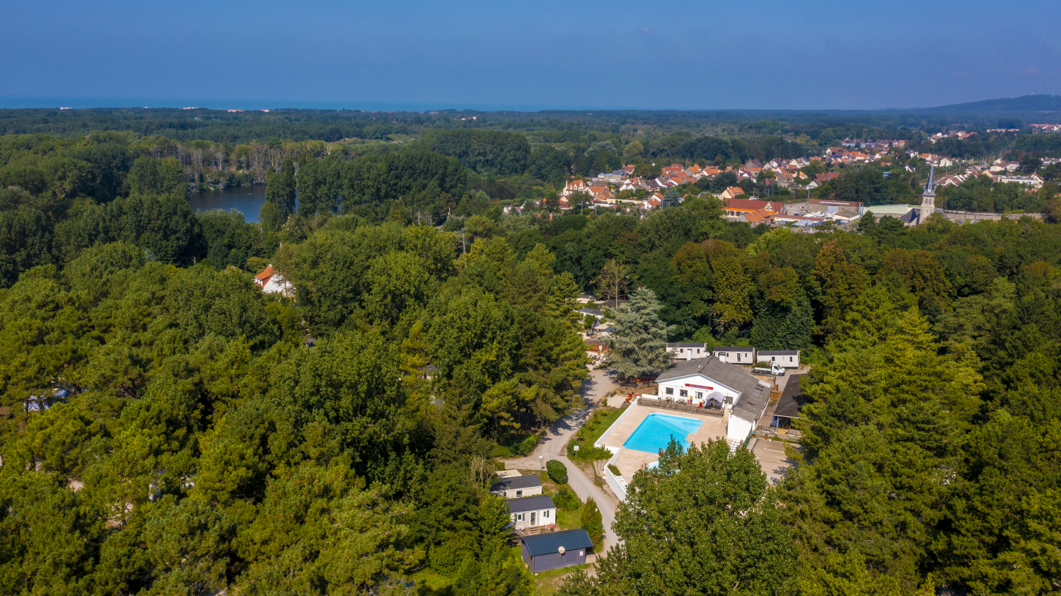 At La Dune Blanche, a spacious outdoor swimming pool is encircled by trees and structures close to a quaint town.