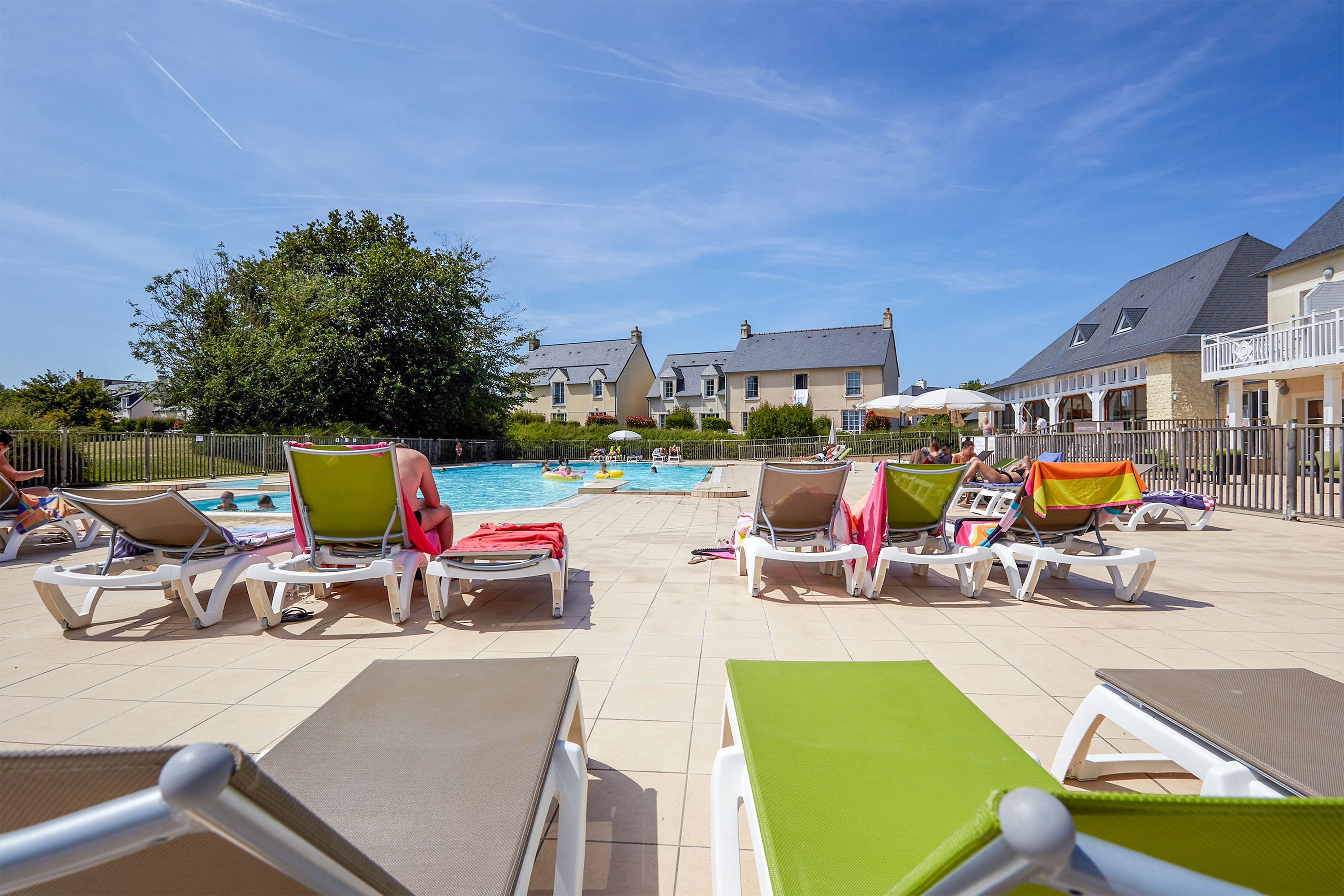 Sunbathers relax on loungers beside a clear swimming pool, surrounded by residential cottages under a bright blue sky at Le Green Beach
