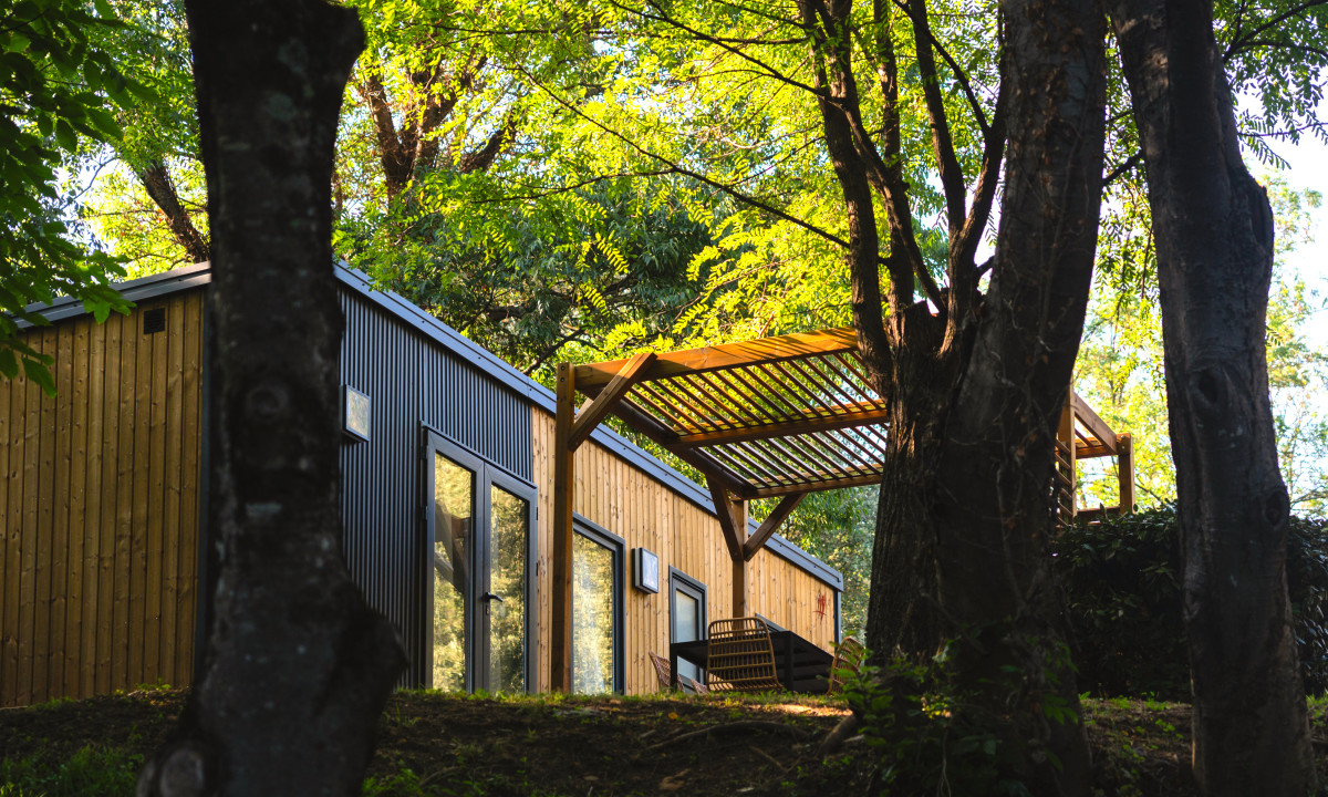 At La Clémentine, a cabin tucked away among trees, sheltered by a wooden arbor, in a sunlit woodland environment.