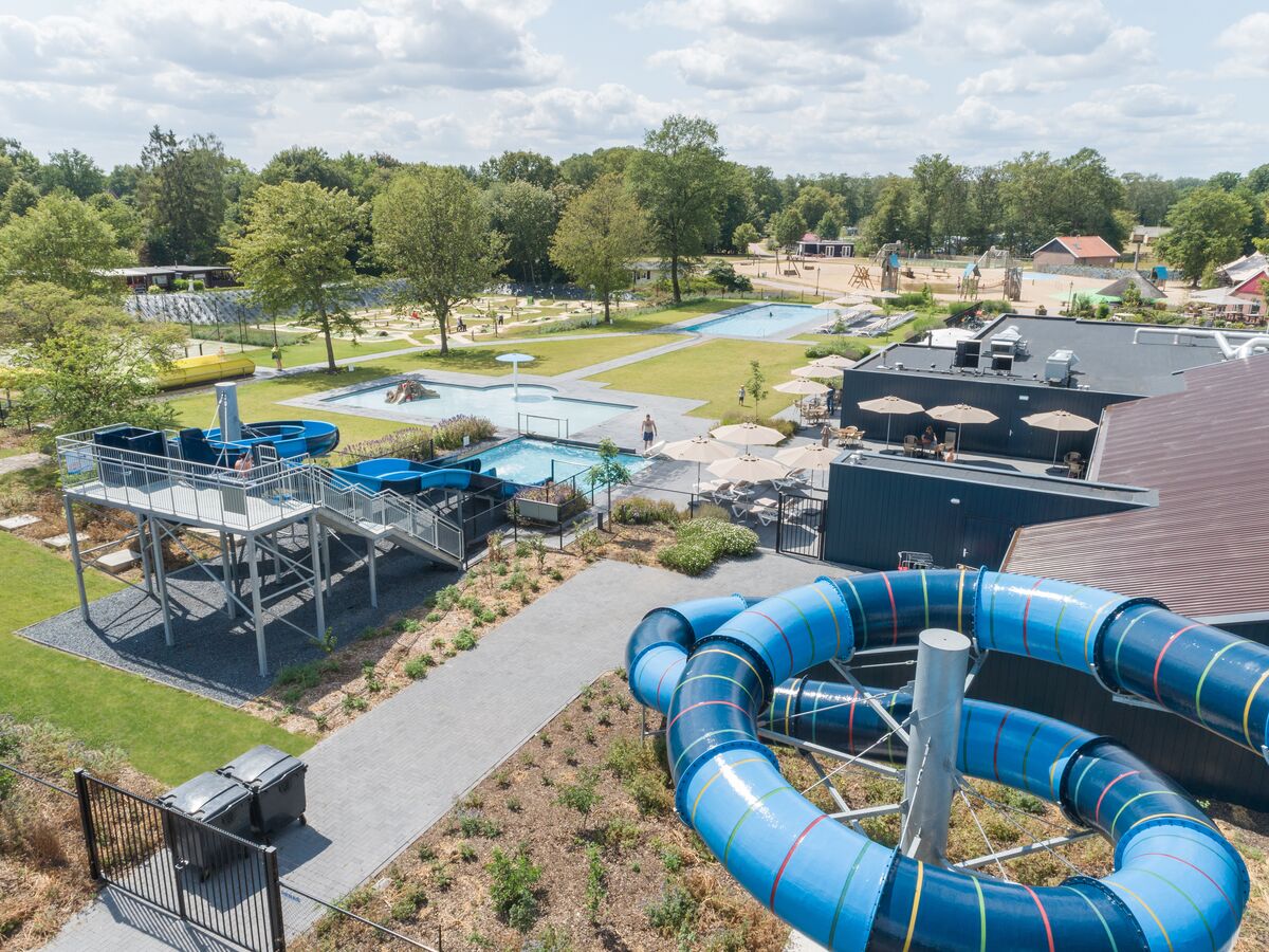 At De Twee Bruggen, a cerulean water slide spirals down into a swimming pool, encircled by verdant lawns, trees, and poolside seating.