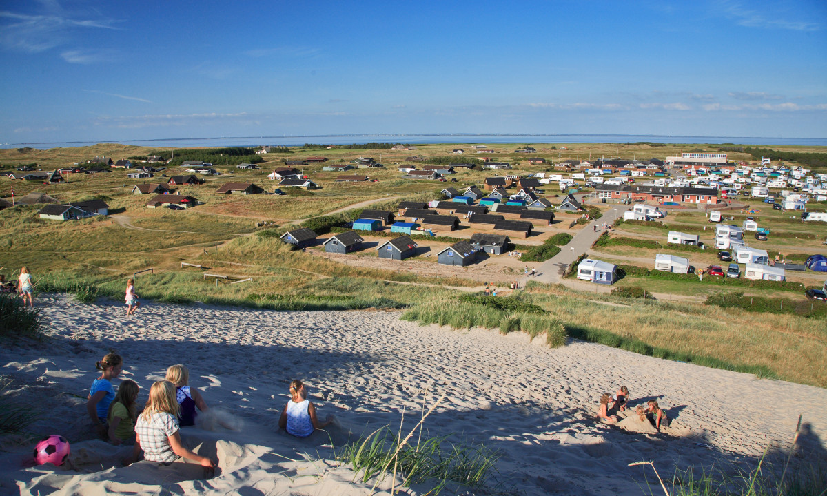 At Dancamps Nordsø, kids frolic on sandy hills, gazing at an extensive campground filled with cabins and trailers beneath a bright blue sky.