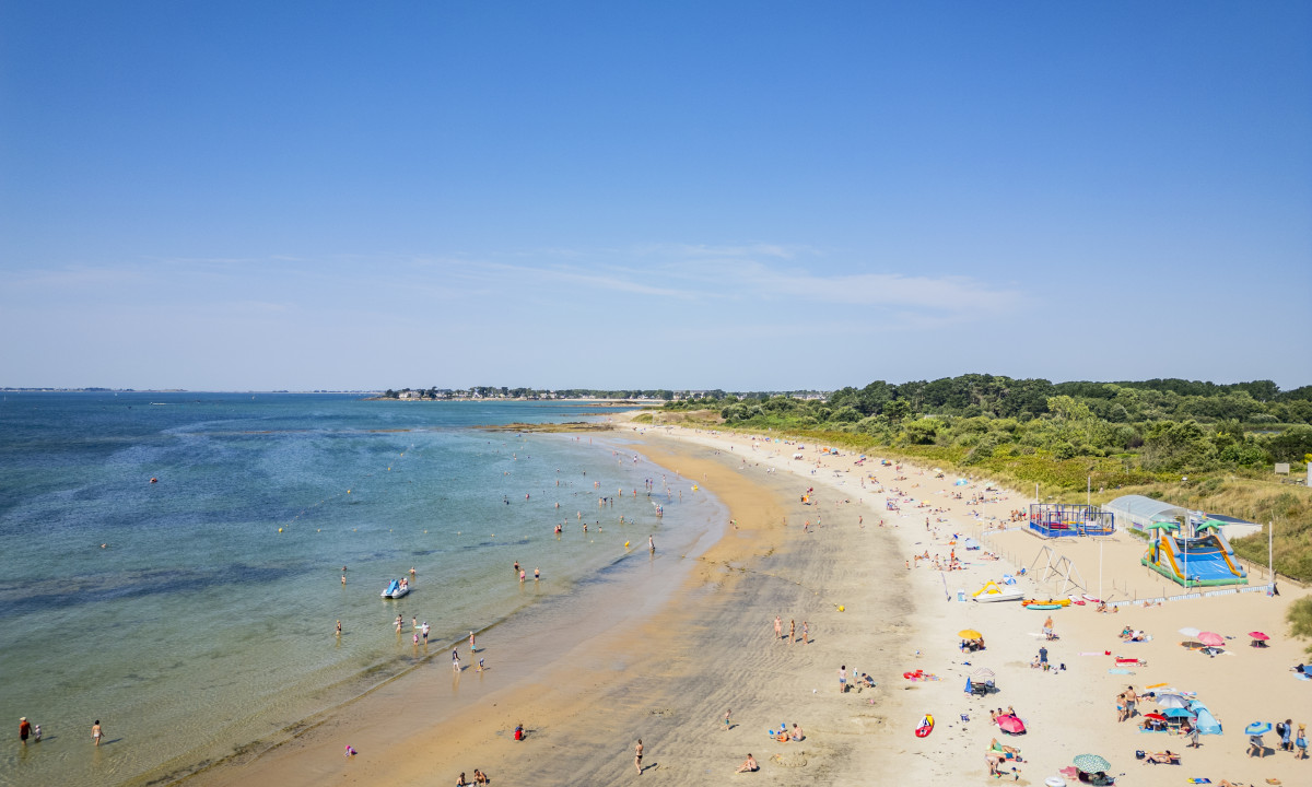 At De la Plage, visitors unwind on the sandy beach, bathe in the ocean, and appreciate vibrant umbrellas beneath a clear azure sky.