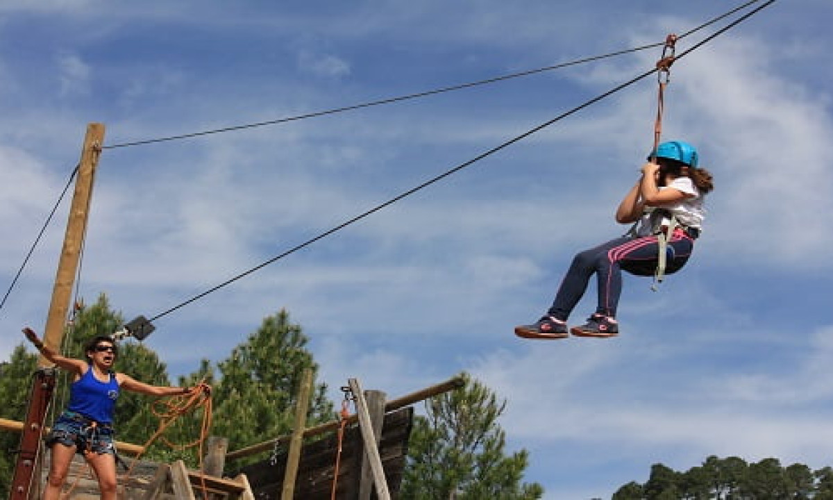 At Puente de las Herrerías, a child is gliding on a zip line while an instructor observes, encircled by trees and a clear sky.