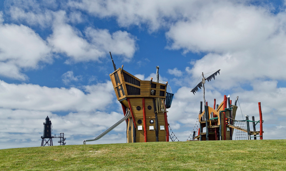 At KNAUS Campingpark Dorum, a wooden play structure designed like a ship sits on lush green grass beneath a blue sky dotted with fluffy clouds.
