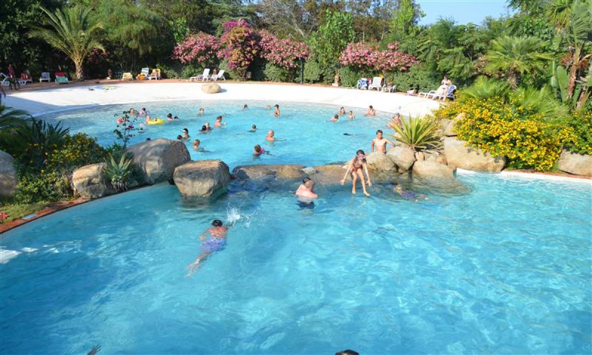 At Ultima Spiaggia, individuals are swimming and relaxing in a spacious outdoor pool encircled by vibrant foliage and colorful blossoms.