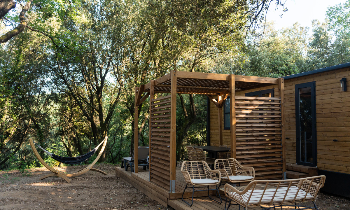 At La Clémentine, a timber deck features wicker seating beneath a pergola adjacent to a cabin, encircled by trees with a hammock nearby.