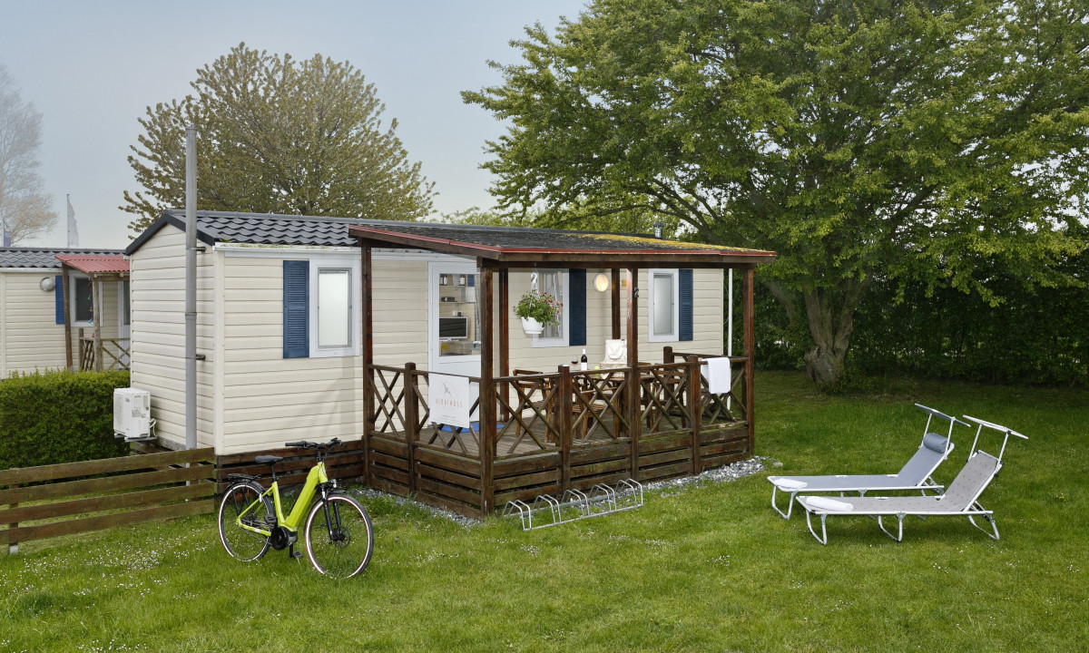 At KNAUS Campingpark Rügen, a mobile home features a sheltered porch and a suspended plant, encircled by grass, trees, a bicycle, and reclining chairs.