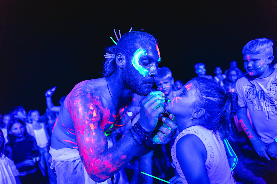 Face-painted man using a brush to paint young girl's face, surrounded by others in neon paint at Residence Village