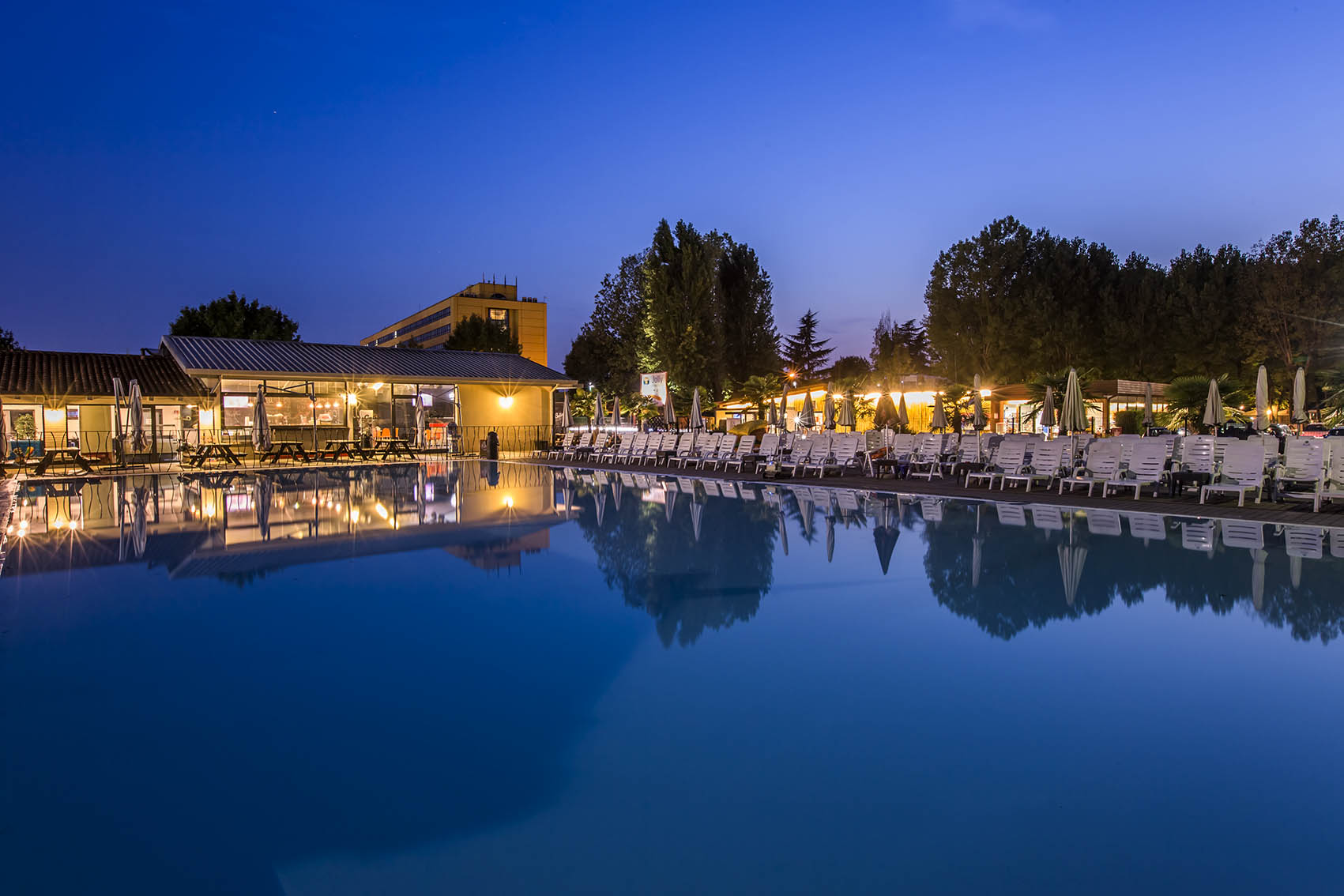 Swimming pool reflecting nearby buildings and trees at dusk, surrounded by many empty lounge chairs at hu Venezia Camping in Town