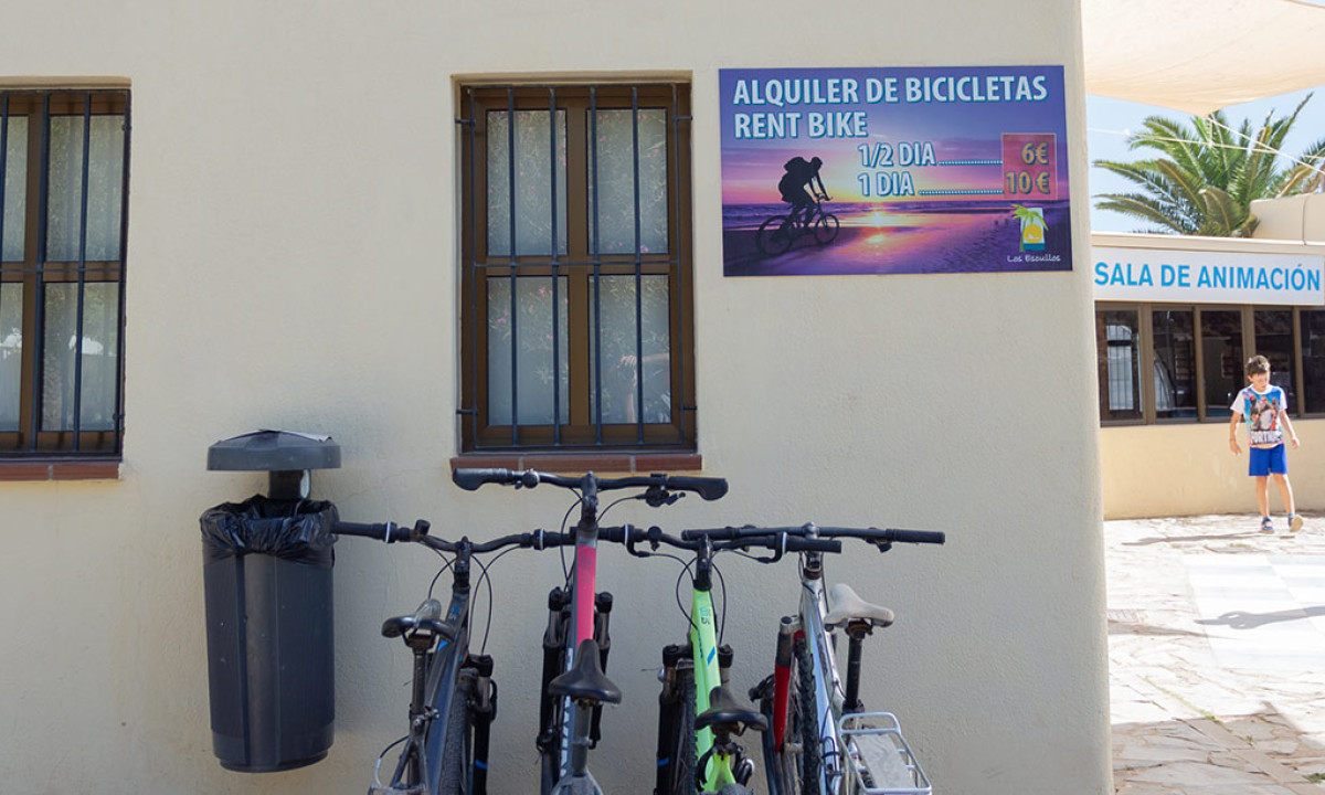At Camping Los Escullos, bicycles are leaned against a tan wall featuring barred windows and a bike rental sign.