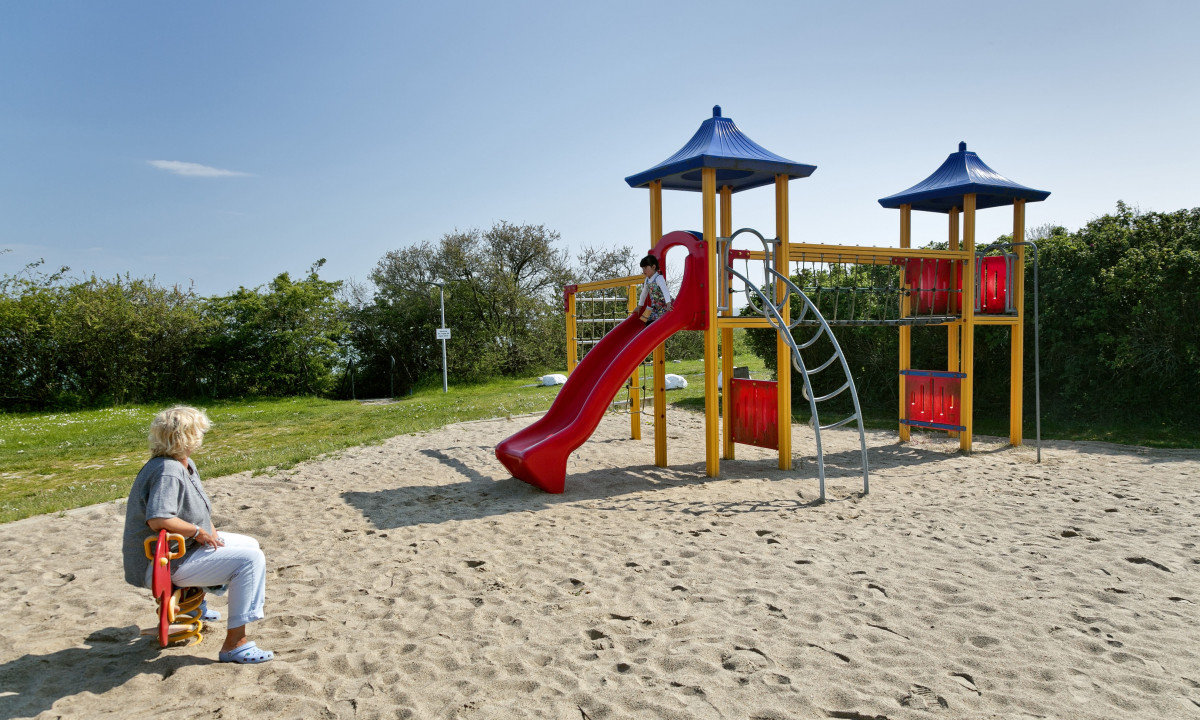 At KNAUS Campingpark Rügen, a playground setup featuring a slide and climbing bars is being utilized by a child, while an elderly woman sits nearby in a grassy area surrounded by trees.