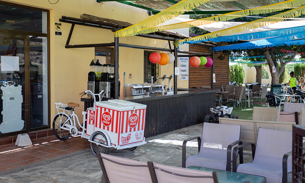 At Camping Los Escullos, there is a seating area adorned with vibrant decorations and a stationary ice cream cart next to an outdoor bar.