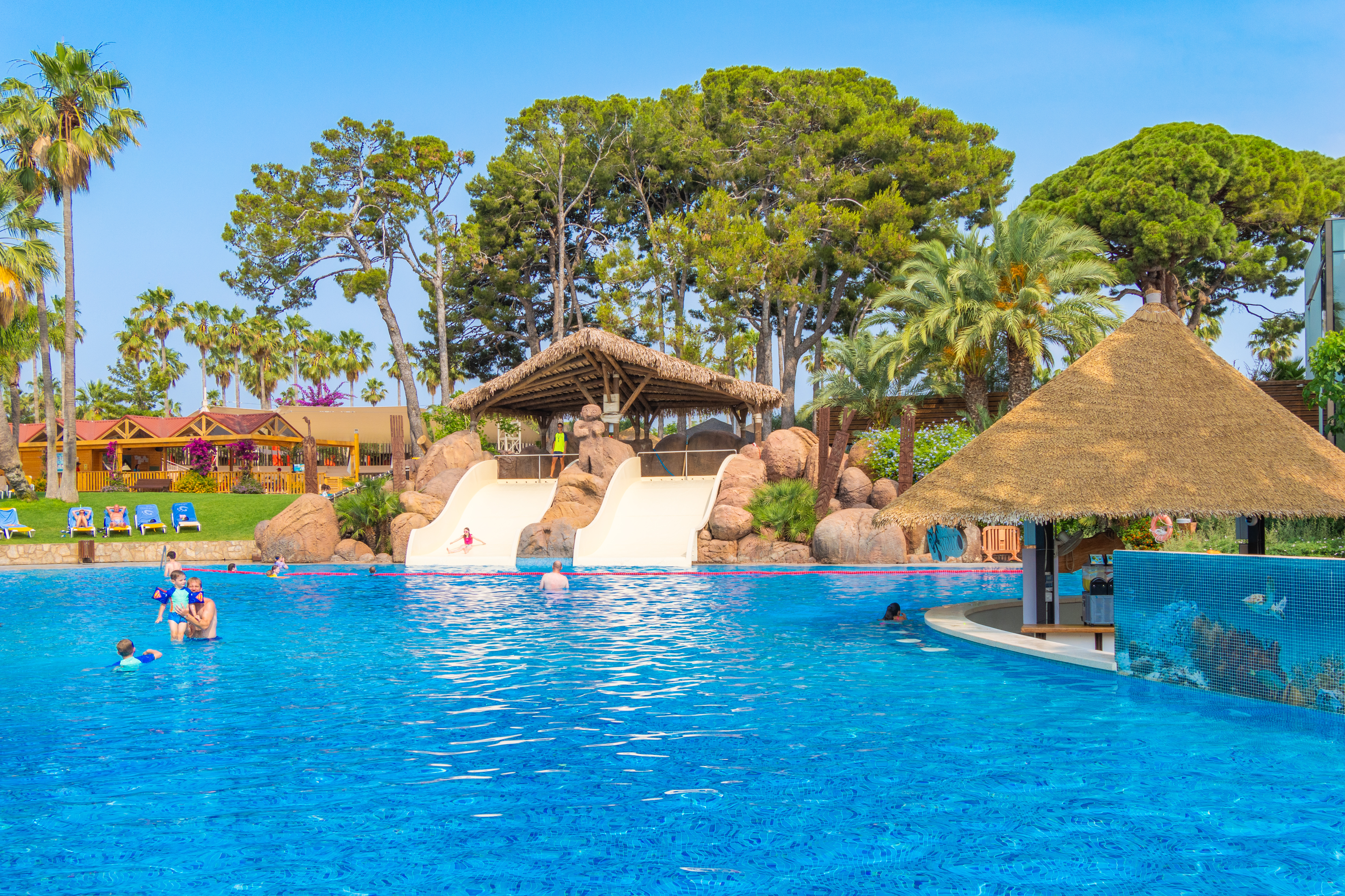 At Cambrils Park, kids are enjoying themselves in a large swimming pool featuring water slides, encircled by trees and sun loungers.