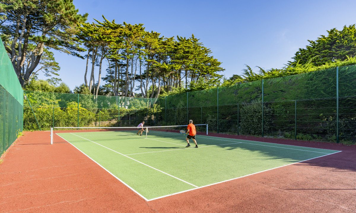 At De la Plage, two individuals are playing tennis on a court encircled by towering trees beneath a clear azure sky.