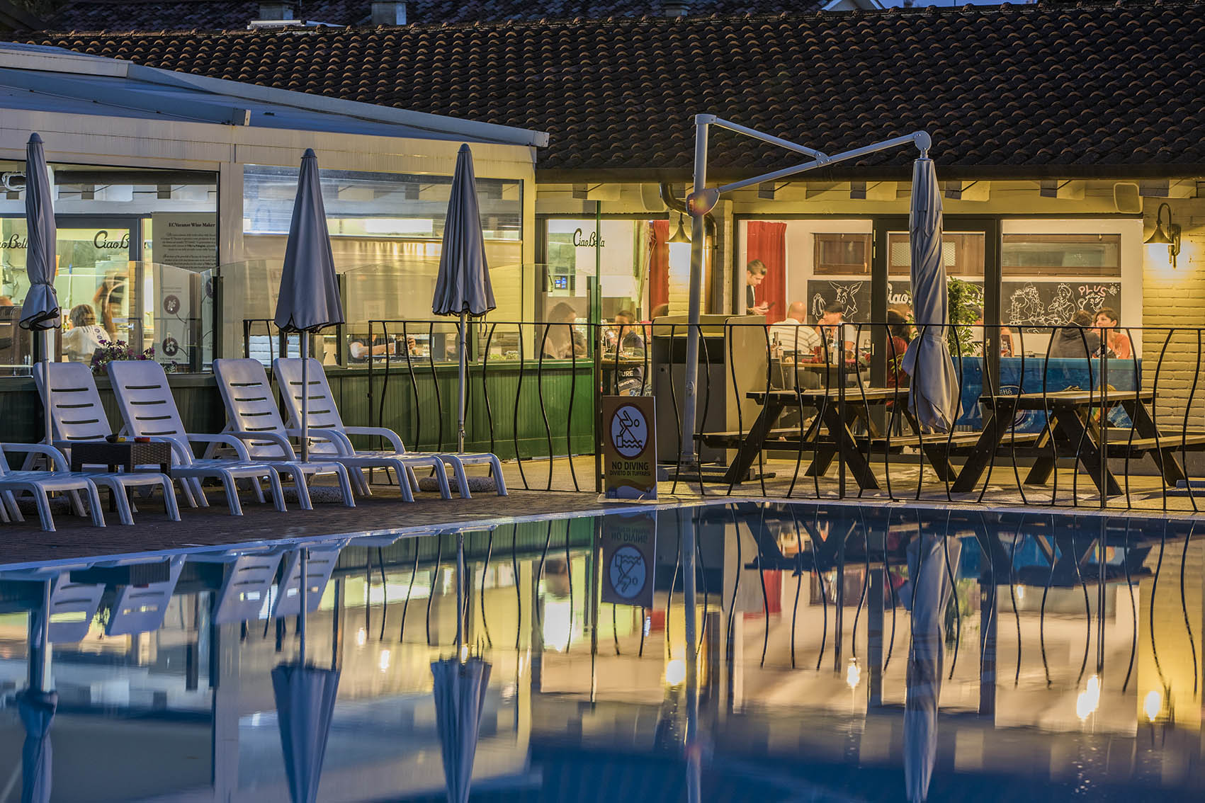 Poolside chairs and umbrellas reflecting in a calm pool surface; behind them, a café with people dining indoors at hu Venezia Camping in Town