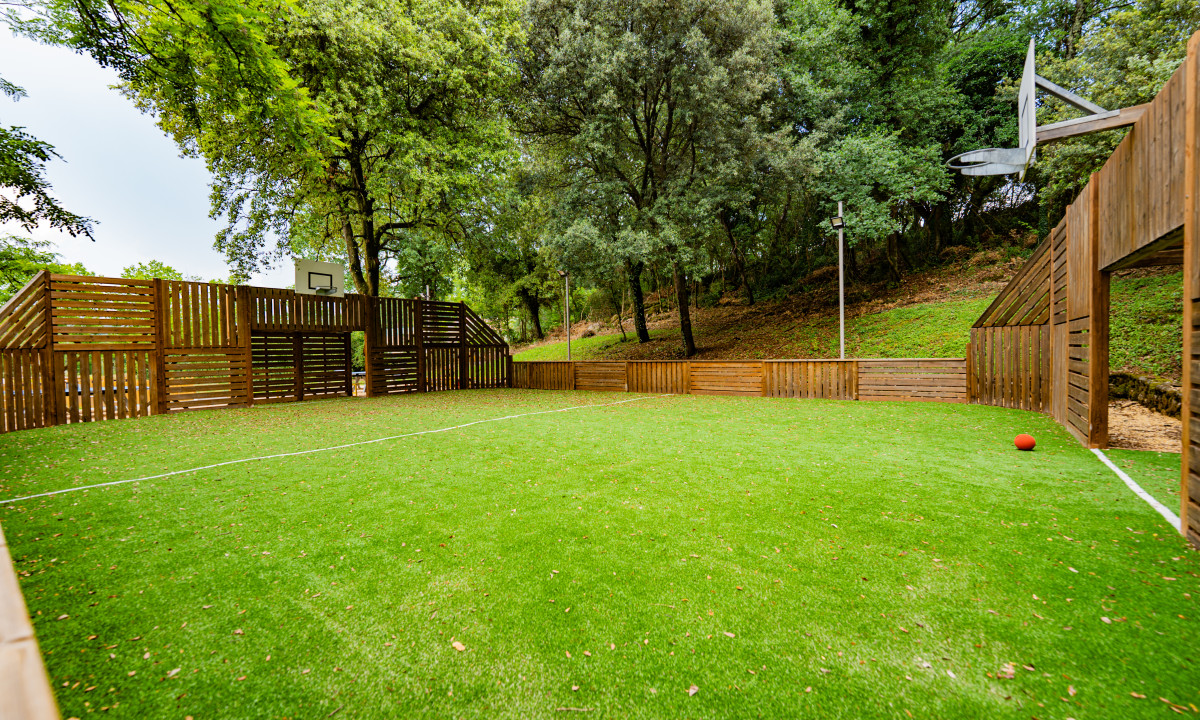 At La Clémentine, a basketball court enclosed by wooden barriers is located in a verdant, tree-filled park, with a red ball nearby.