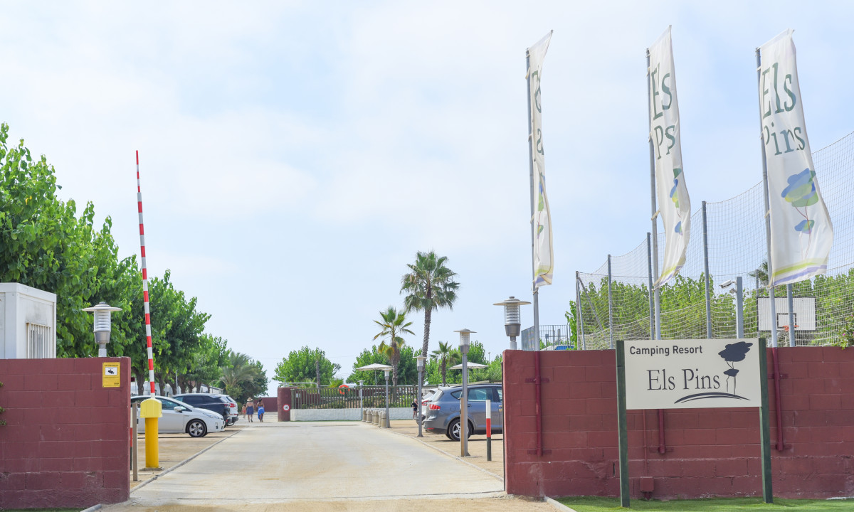 Entrance gate with a barrier arm and flags fluttering, surrounded by trees and parked cars at Resort Els Pins