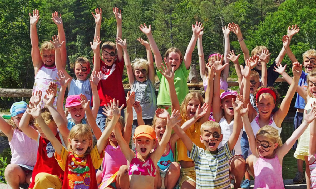 At Les Ranchisses, kids with decorated faces are joyfully raising their hands in front of a wooden fence and a forest.