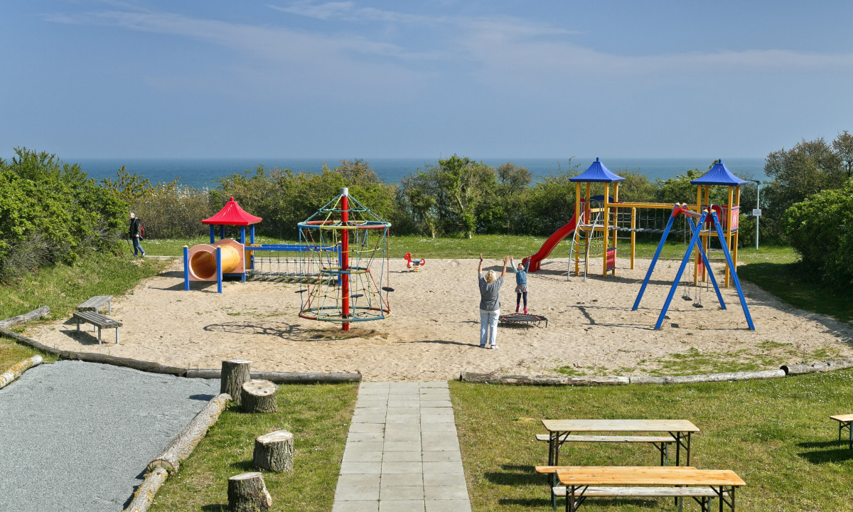 At KNAUS Campingpark Rügen, there is a seaside playground equipped with slides, swings, and climbing apparatus; kids are playing while adults oversee them in the grassy area.