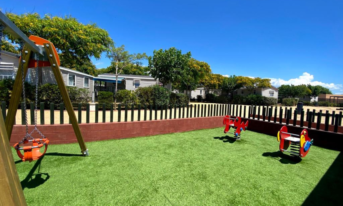 Swing set and see-saws in enclosed play area with green turf, surrounded by trees and mobile homes at Resort Els Pins