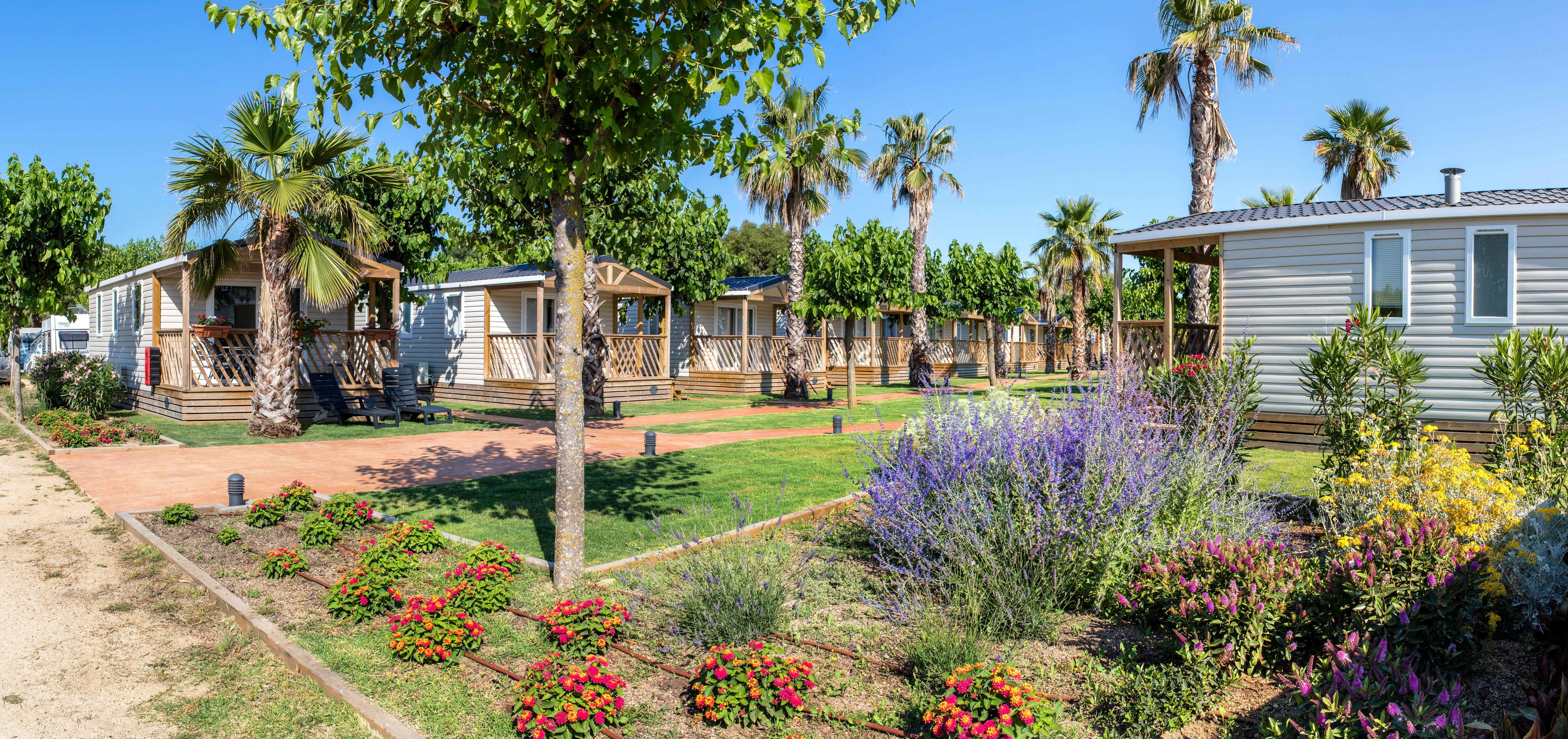 Mobile homes with porches among trees and colorful flower beds; clear blue sky at El Pinar