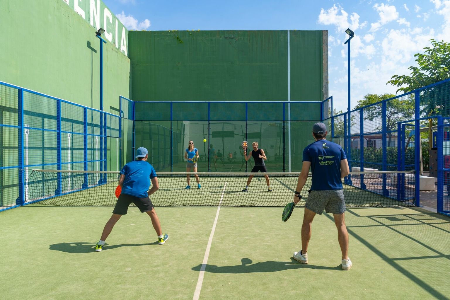 At Valencia, four individuals engage in paddle tennis on a court surrounded by green walls and a clear blue sky.