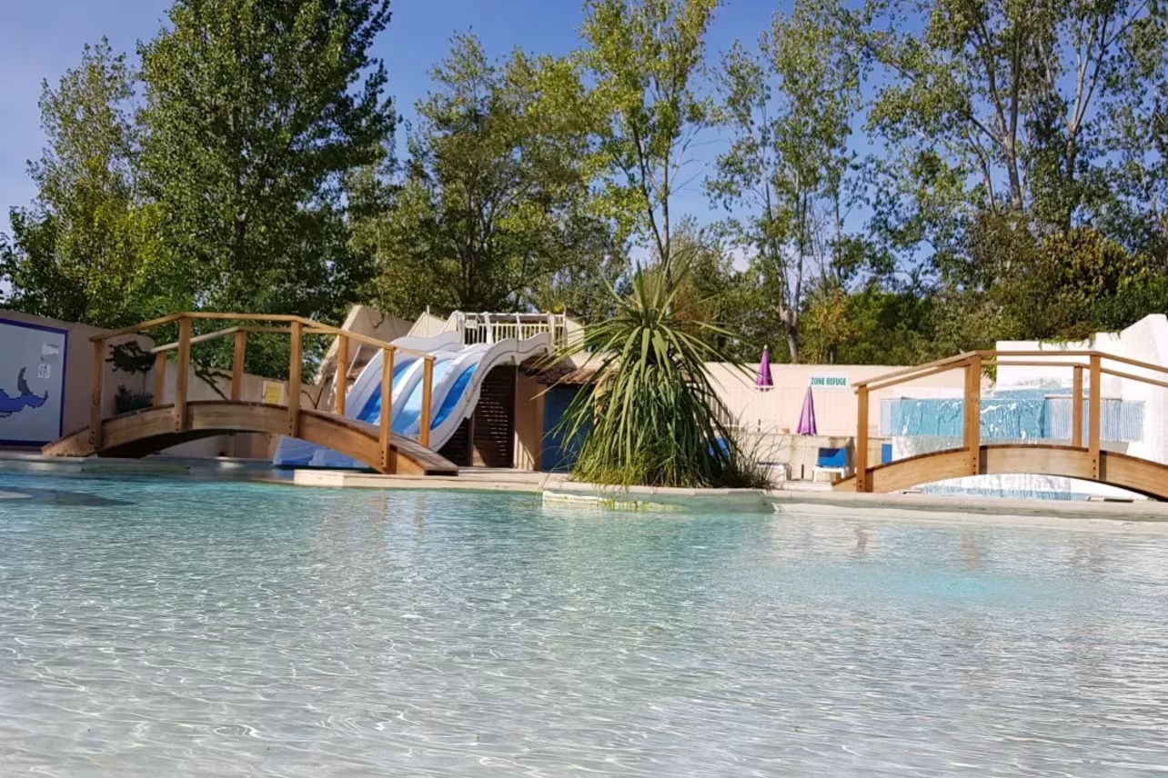 At Les Jardins de Tivoli, there is a waterslide adjacent to a wooden bridge, overlooking a pool with trees in the background.