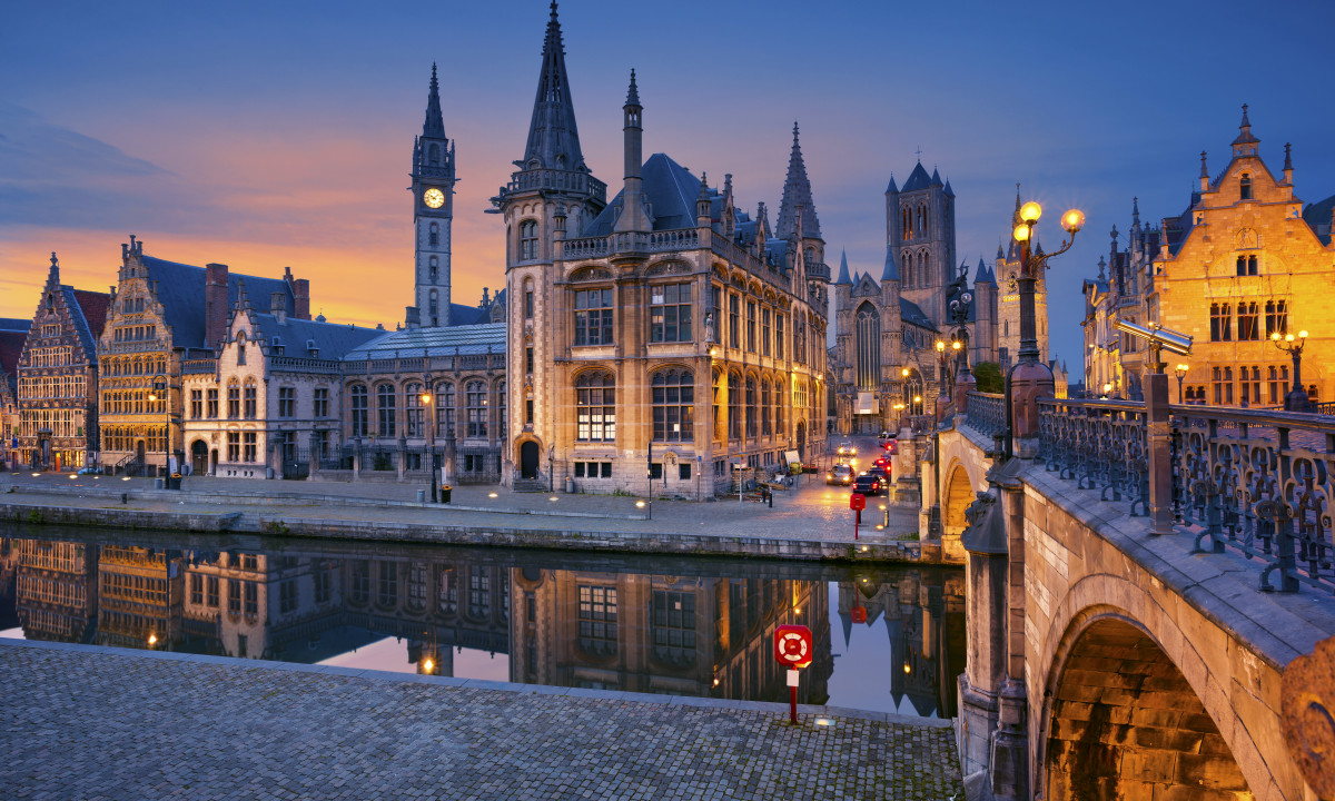 At Urban Gardens Gent, a bridge spans the canal, mirroring the lit Gothic structures at twilight, featuring a clock tower and streetlights in a historic cityscape.