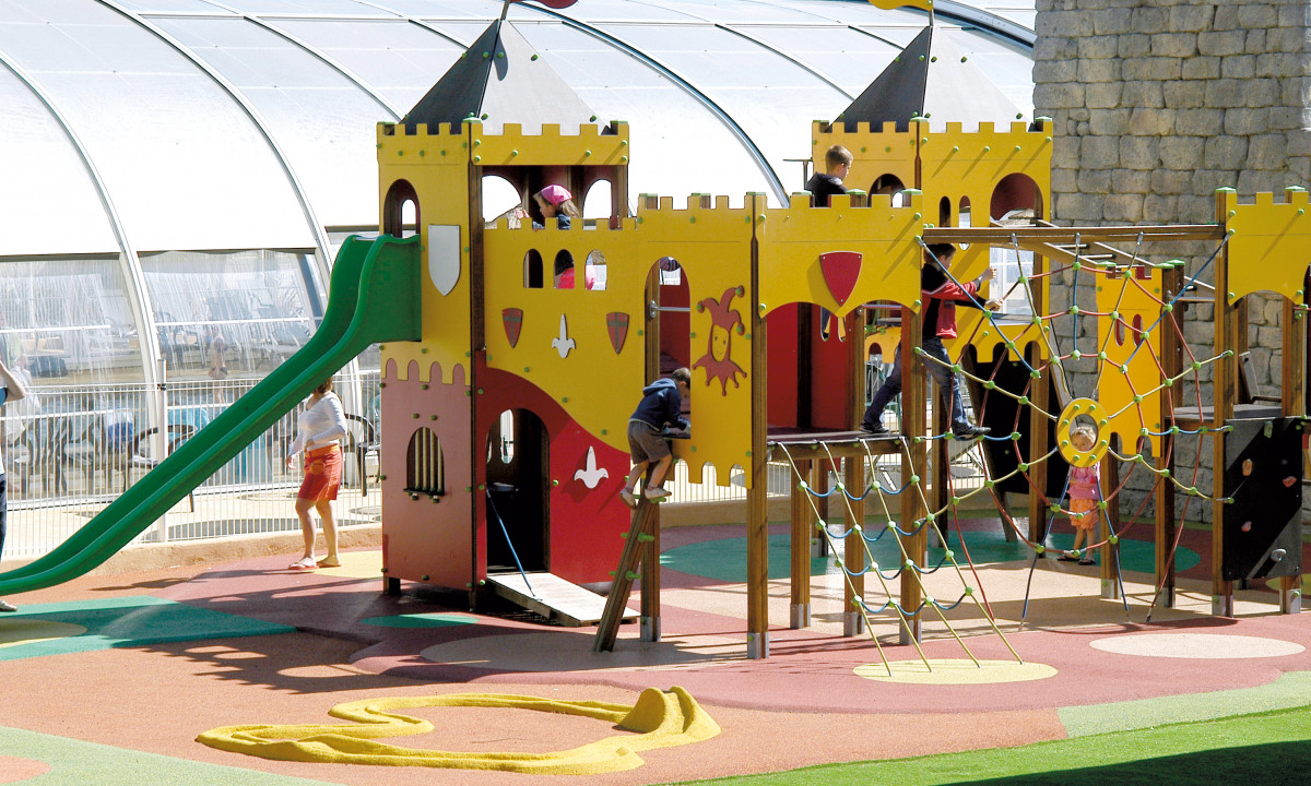 At Le Domaine de Léveno, children are enjoying themselves on a vibrant castle-themed play structure, featuring slides and climbing nets, situated close to a domed enclosure.