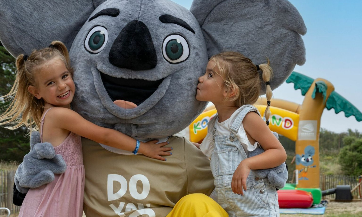 Two smiling children embrace a large koala mascot, set in a playful outdoor area with inflatable structures at Maguide