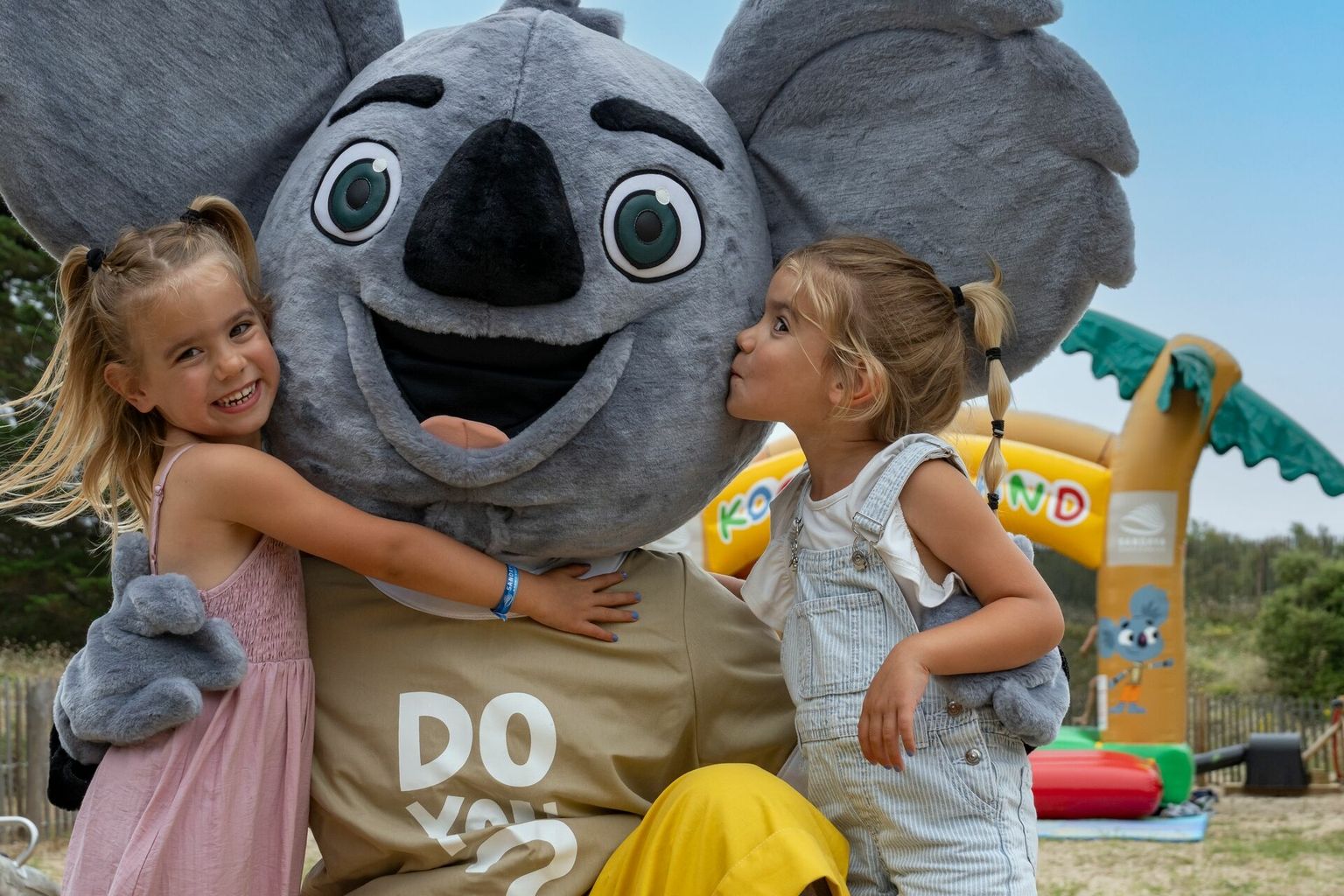 Two smiling children embrace a large koala mascot, set in a playful outdoor area with inflatable structures at Maguide
