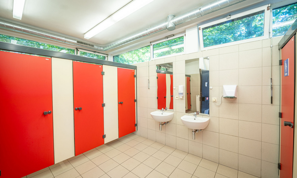 At Urban Gardens Gent, there are basins and commodes in a tidy, tiled restroom featuring crimson stall doors and fluorescent illumination.