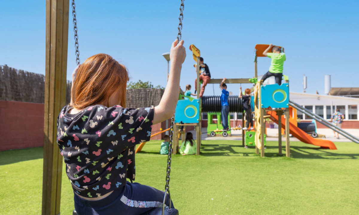 Child swings alone, observing distant playground equipment with other children playing. Sunny day in an outdoor recreational area at Resort Els Pins