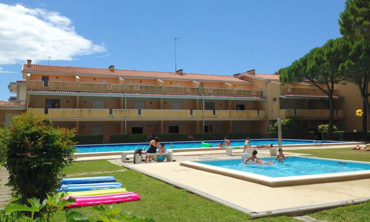 People swim and sunbathe around outdoor pools in front of a multi-story building, surrounded by grass and trees at Villaggio Selene