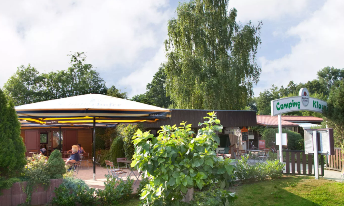 At KNAUS Campingpark Lübben, individuals are seated beneath a large parasol outside a timber structure, encircled by lush vegetation.