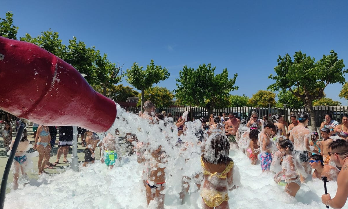 Large foam cannon spraying bubbles on a crowd of people in swimwear outdoors, surrounded by trees and fences at Resort Els Pins