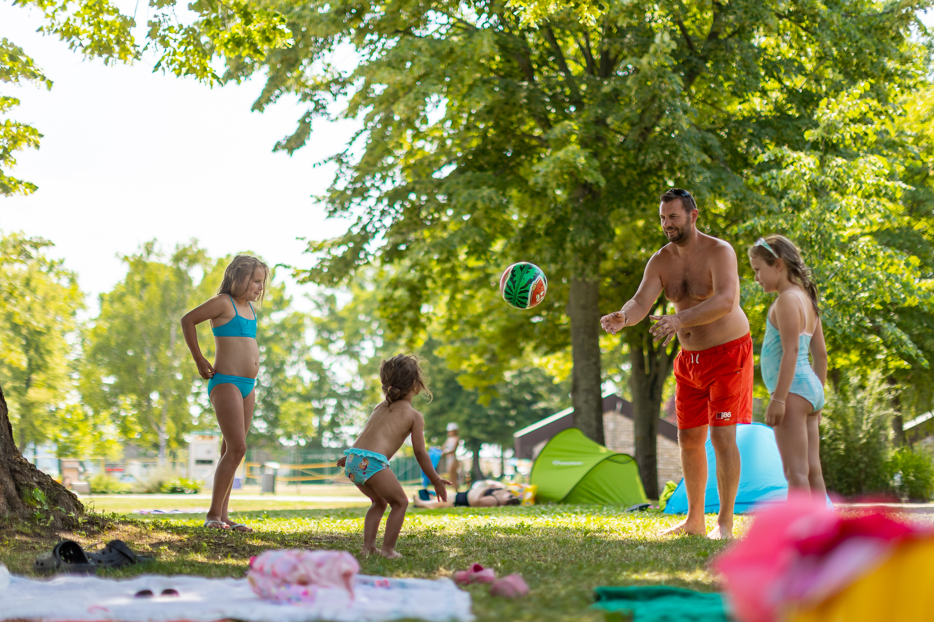 Children play ball with an adult amidst green trees and tents at Füred