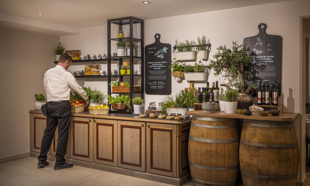 A man arranges baskets of fruits and vegetables on a countertop surrounded by plants and bottles, indoors at Valamar Obertauern Hotel
