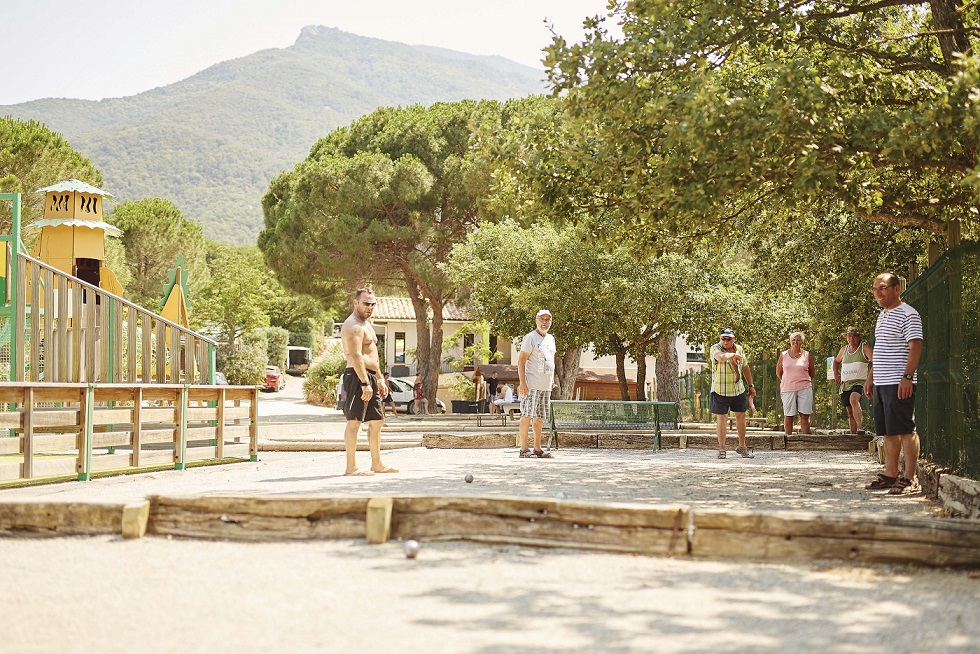 Group of people playing pétanque on a gravel court, surrounded by trees and a playground at Les Albères