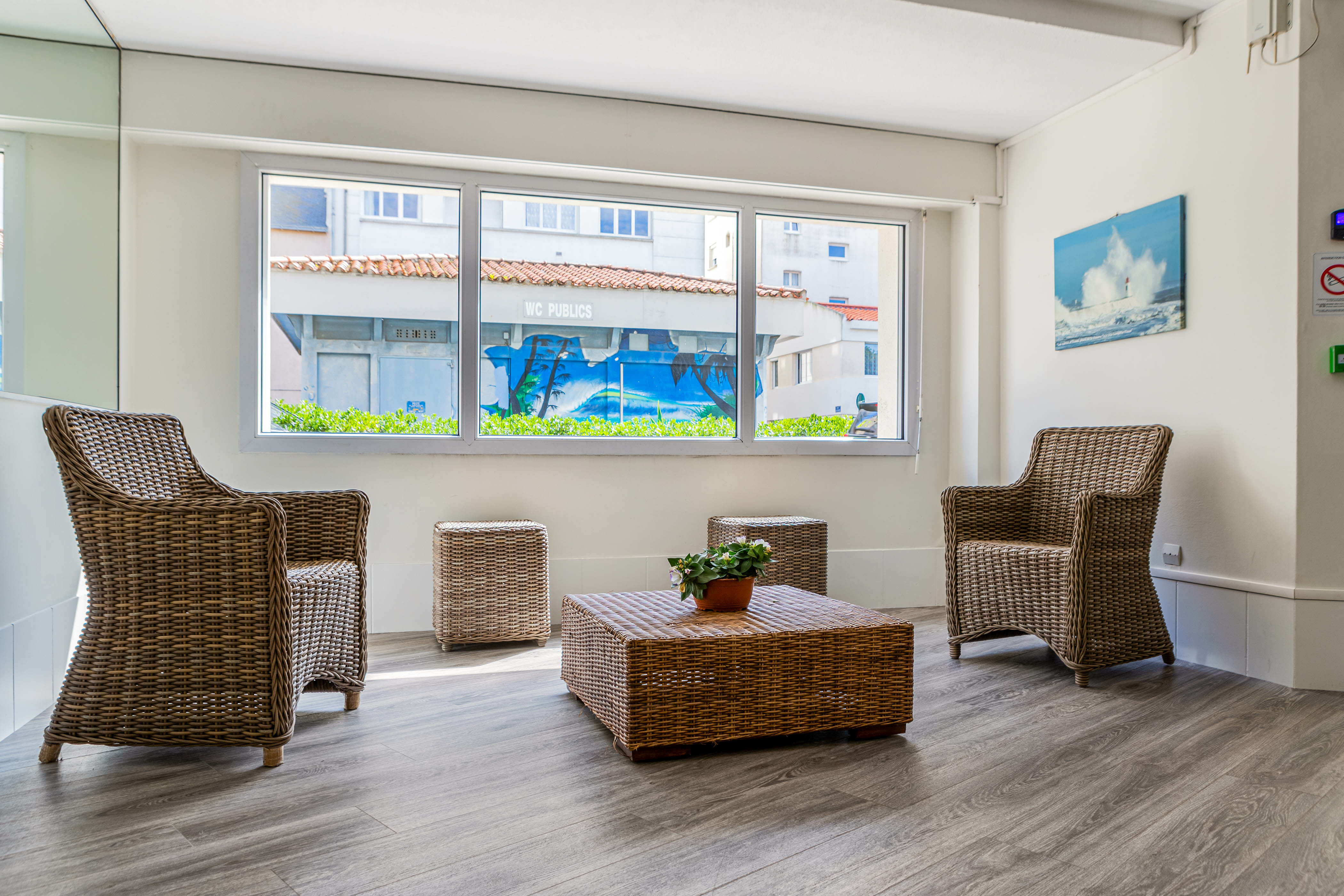 Wicker chairs and table sit in a sunlit room with large windows, overlooking an outdoor mural of a beach scene at La Baie des Sables